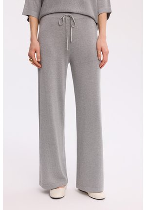 Szare szerokie dzianinowe spodnie z troczkiem w talii noszone z białymi butami oraz część pasującego swetra widoczna na jednolitym tle.