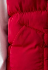 Gros plan d'une personne portant une veste matelassée rouge avec une poche latérale et une chemise à manches longues de couleur claire en dessous.
