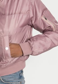Różowa satynowa kurtka bomberka z ściągaczami przy mankietach i pasie. Posiada kieszeń na suwak na rękawie oraz kieszenie boczne. Gładka faktura i dopasowany krój.