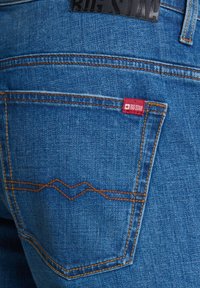 Mėlynos džinsinės kelnės su oranžiniu zigzago siūlų raštu ant galinio kišenės, su raudonu audinio užrašu "BIG STAR." Tekstūruoto audinio artimas vaizdas.