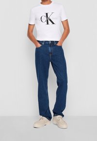 Osoba ubrana w białą koszulkę z logo Calvin Klein Jeans, niebieskie dżinsy o prostym kroju i beżowe buty na sznurówki, stoi z rękami w kieszeniach.