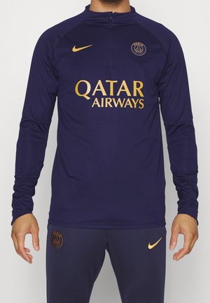 Uomo che indossa abbigliamento sportivo Paris Saint-Germain blu navy con loghi Nike dorati e testo dello sponsor Qatar Airways sulla maglietta.