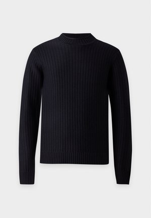 Homme debout, les mains dans les poches, portant un pull noir côtelé, un pantalon cargo gris et des chaussures noires sur fond blanc.