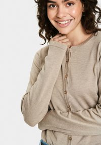 Bēša garenā piedurknē kardigāns ar ribotām tekstūrām, kas aprīkots ar piecām brūnām pogām priekšā un locītām cuffs.