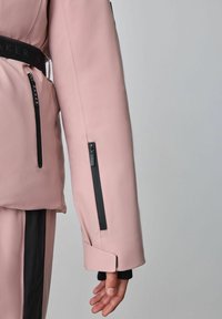 Lietusmētelis rozā krāsā ar gludu struktūru, ar melniem rāvējslēdzējiem un akcentiem. Ietver jostu jostasvietā un ciešus aproces uz piedurknēm.