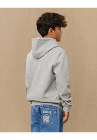 Felpa grigia con cappuccio a coulisse, polsini e fondo a coste, abbinata a jeans blu con logo di Yale e tasca con motivo.