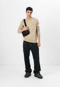 Beige pamut pulóver bordás mandzsettákkal, fekete keresztpántos táska, fekete laza szabású farmer és fekete textúrázott sportcipő.