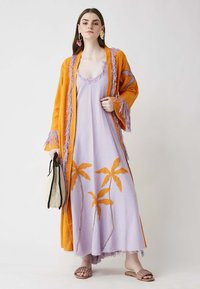 Kimono de flecos naranjas sobre un vestido lavanda con bordado de hojas naranjas, combinado con sandalias planas y un bolso de mimbre. Textura de tela suave.