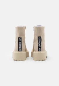 Un paio di stivaletti beige in suede con suole spesse e linguette in tessuto nero che presentano il testo bianco "Steve Madden", visti da dietro.