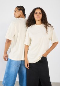 Kremfarget, oversized bomulls-t-skjorte med senkede skuldre, med en rund halsutskjæring. Båret av to modeller med jeans og svarte skjørt.