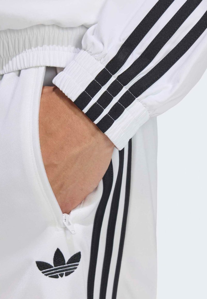 Mano nella tasca dei pantaloni della tuta Adidas bianchi con strisce nere e logo, manica abbinata con strisce nere visibile.