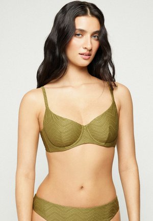 Mujer con cabello largo y oscuro que lleva un sujetador verde oliva texturizado y ropa interior a juego, de pie frente a un fondo liso y claro.