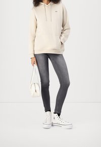 Felpa beige con tasca frontale e logo bianco, abbinata a jeans skinny grigi e sneakers alte bianche. Tiene una piccola borsa bianca.