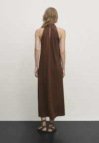Robe maxi marron à dos nu avec un col licou, coupe ample, dotée d'un détail en forme de trou de serrure dans le dos. Texture lisse, tombé élégant jusqu'au sol.