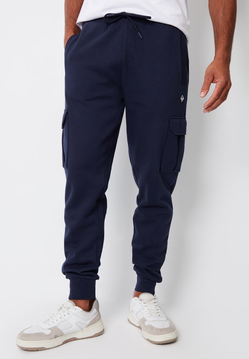 Uomo che indossa jogger cargo blu navy con vita con coulisse e sneakers bianche, in piedi contro uno sfondo chiaro uniforme.