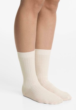 Ben som bär ljusbeige, ribbade strumpor som slutar vid vaden, står på en vit yta med en enkel vit bakgrund.
