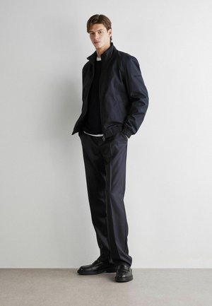 Giovane uomo in piedi che indossa una giacca nera con cerniera, pantaloni neri e scarpe nere contro una parete bianca uniforme.