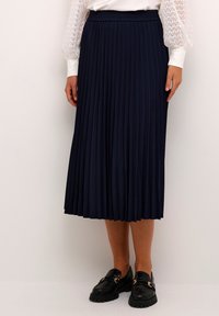 Marinblå plisserad midikjol i mjukt tyg med elastisk midja. Bärs med en vit långärmad skjorta och svarta loafers.