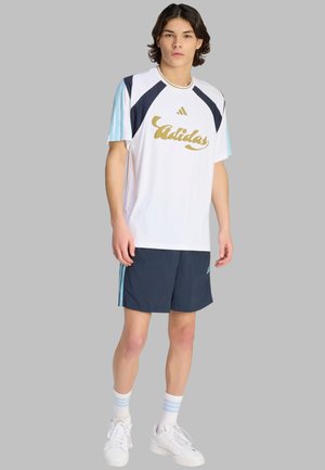 Joven con camiseta blanca de Adidas con logo dorado, pantalones cortos azul marino con rayas azul claro, calcetines blancos y zapatillas blancas sobre fondo gris.