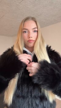 Czarna kurtka ze sztucznego futra z długimi rękawami i miękkim, teksturowanym wykończeniem. Pod spodem noszona jest jasnoszara bluza z kapturem, a model zapina kurtkę.