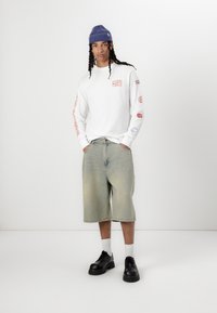 White long-sleeve shirt with colorful graphics, light blue denim shorts, and black lace-up shoes. The model wears a blue knit beanie.