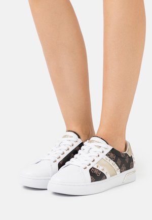 Jambes portant des baskets blanches basses avec des côtés à motifs noir et or et des détails cloutés dorés, présentées sur un fond blanc.