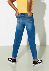Jeans de denim azul con cintura alta, diseño de cinco bolsillos y dobladillo deshilachado. Combinados con zapatillas negras y blancas, y una blusa amarilla texturizada.