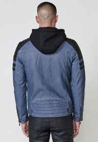 Uomo visto di spalle che indossa una giacca di denim blu con toppe imbottite nere sulle spalle e un cappuccio nero, abbinato a pantaloni neri.
