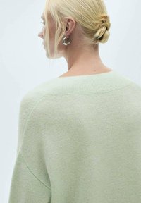 Hellgrüner, oversized Pullover aus einem weichen, strukturierten Stoff, mit weitem Rundhalsausschnitt und tiefen Schultern. Runde silberne Ohrringe.