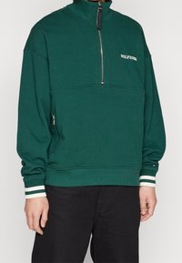 Sudadera verde con cuello alto, cierre delantero de media cremallera y puños a rayas blancas. Cuenta con un logotipo bordado "HILFIGER" y un bolsillo lateral con cremallera.
