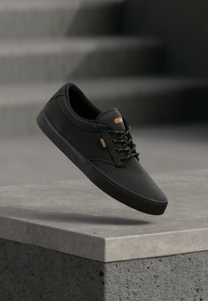 Sorte lærret sneakers med teksturert overflate, sort gummisåle, brune snørebånd og en liten oransje logo på siden.