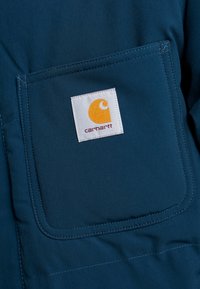 Primer plano de un bolsillo de prenda azul que presenta una etiqueta blanca cosida con un logo naranja y texto "carhartt" en rojo.