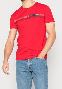 Rød bomulds t-shirt med rund halsudskæring og korte ærmer, med et sort logo og vandrette striber over brystet. Bæres med blå denim jeans.