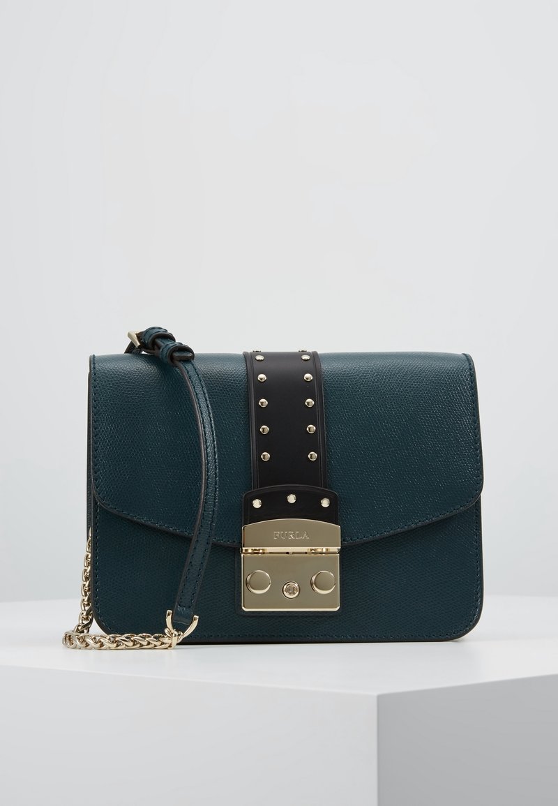 Borsa a tracolla in pelle testurizzata verde scuro con tracolla nera con borchie e dettagli in catena e chiusura dorata su superficie bianca.