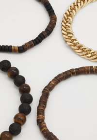 Fire smykker med varierte materialer: brune treperler, svarte lava-perler og en gullkjede. Hver av dem viser unike former og teksturer.