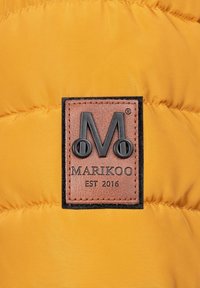 Geel pufferjack met een gestikt patroon. Beschikt over een rechthoekig leren label met een logo en de merknaam "MARIKOO EST 2016" in reliëftekst.