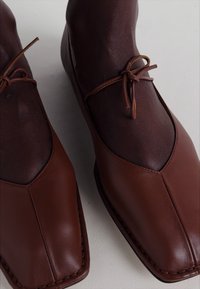 Chaussures en cuir brun avec un bout carré arrondi, présentant un dessus lisse et un lien ajustable et fin au niveau de la cheville.