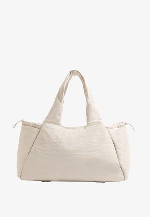 Bézs színű, steppelt táska két füllel, kerek alakú, texturált anyagból készült, és egy zsinóros oldalzsebbel rendelkezik. Simább felület és minimalista dizájn jellemzi.