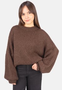 Brauner übergroßer Strickpullover mit strukturierter Oberfläche, weiten Ärmeln und Rundhalsausschnitt, kombiniert mit schwarzen, eng anliegenden Hosen.