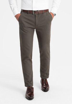 Pantalones verde oliva oscuro con una textura suave, corte recto y bolsillos inclinados. Combinados con zapatos de cuero marrón y un cinturón marrón.