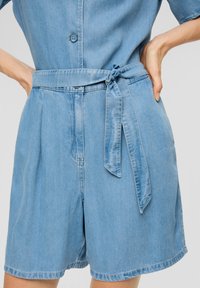 Combinaison en denim bleu clair, à manches courtes, avec une ceinture nouée à la taille et une fermeture à boutons à l'arrière. Tissu texturé avec une finition lisse.