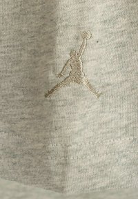 Grau, strukturiertes Stoff-T-Shirt mit kleinem, gesticktem Jumpman-Logo in Gold auf der Vorderseite, nahe dem Saum.