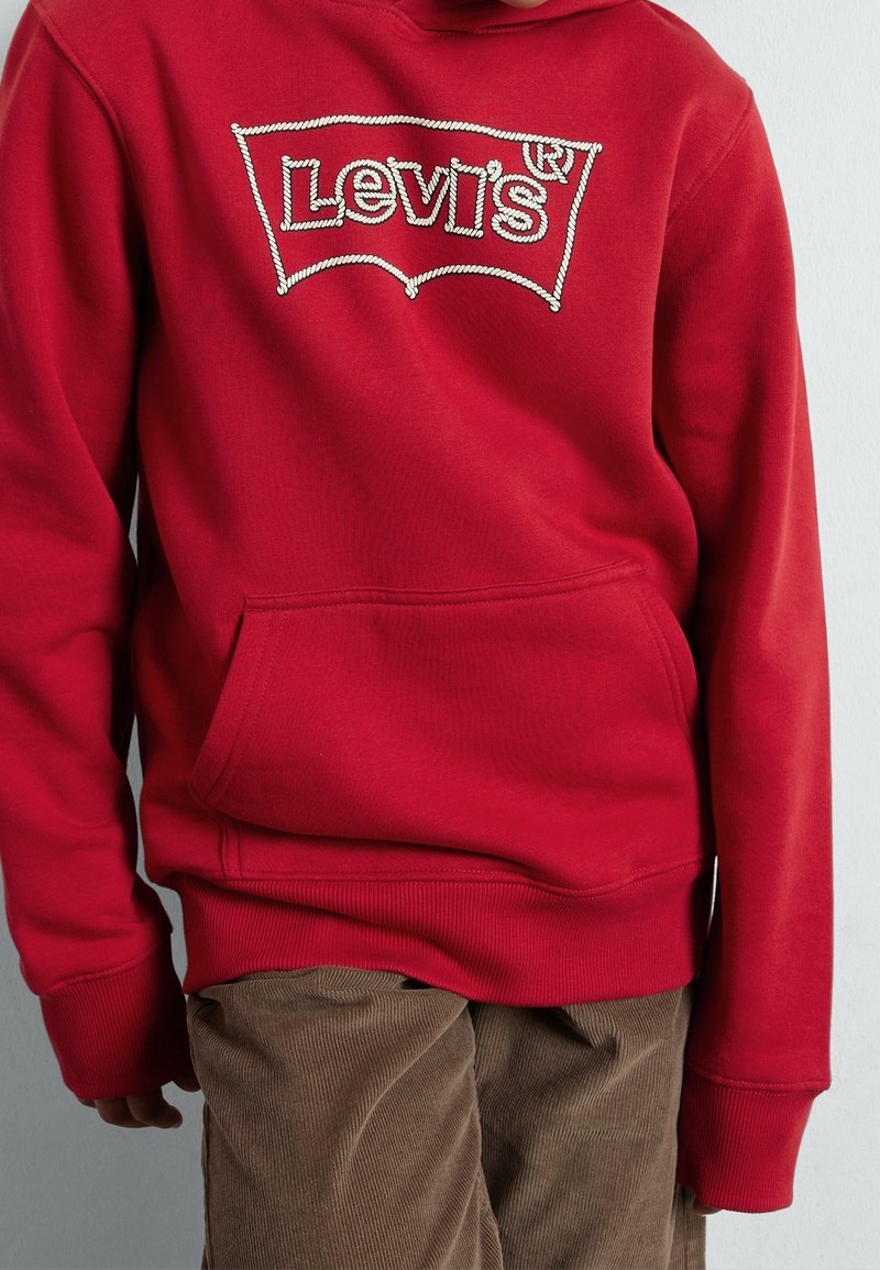 Enfant portant un sweat à capuche rouge Levi’s avec poche avant et pantalon en velours côtelé marron, debout devant un fond clair uni.