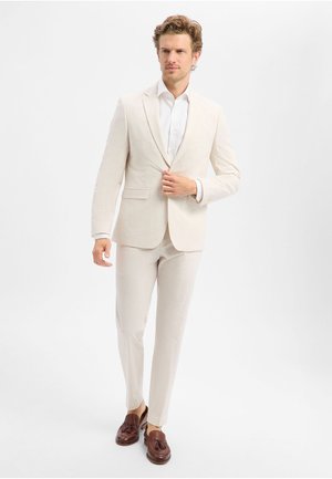 Man in beige pak en wit overhemd die zijn jasje afstelt, lopend, draagt bruine loafers met kwastjes, tegen een effen lichte achtergrond.