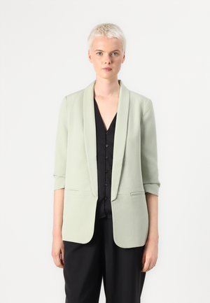Světlý zelený blazer s prošívaným límcem, rukávem ke třičtvrtinám a předními kapsami, vrstvený přes černou košili na knoflíky. Hladká textura.