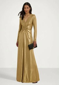 Robe dorée à manches longues avec un détail frontal tordu, confectionnée dans un tissu brillant. Les accessoires comprennent une petite pochette noire avec un fermoir doré.