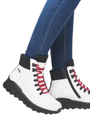 Bottines blanches avec des accents noirs, lacets à motifs rouges, fermeture éclair sur le côté et semelle noire texturée. Portées avec des leggings en denim bleu.
