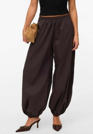 Kvinde iført løstsiddende mørkebrune elastikbukser med rynkede kanter, sort ærmeløs top, brune højhælede mules, holder en tan-farvet clutch.