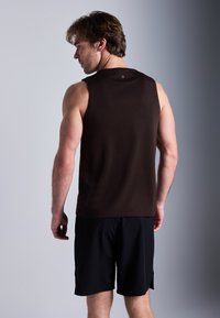 Camisola castanha sem mangas com uma textura suave, combinada com calções pretos. A camisola tem um decote redondo e uma etiqueta com o logótipo na parte de trás.