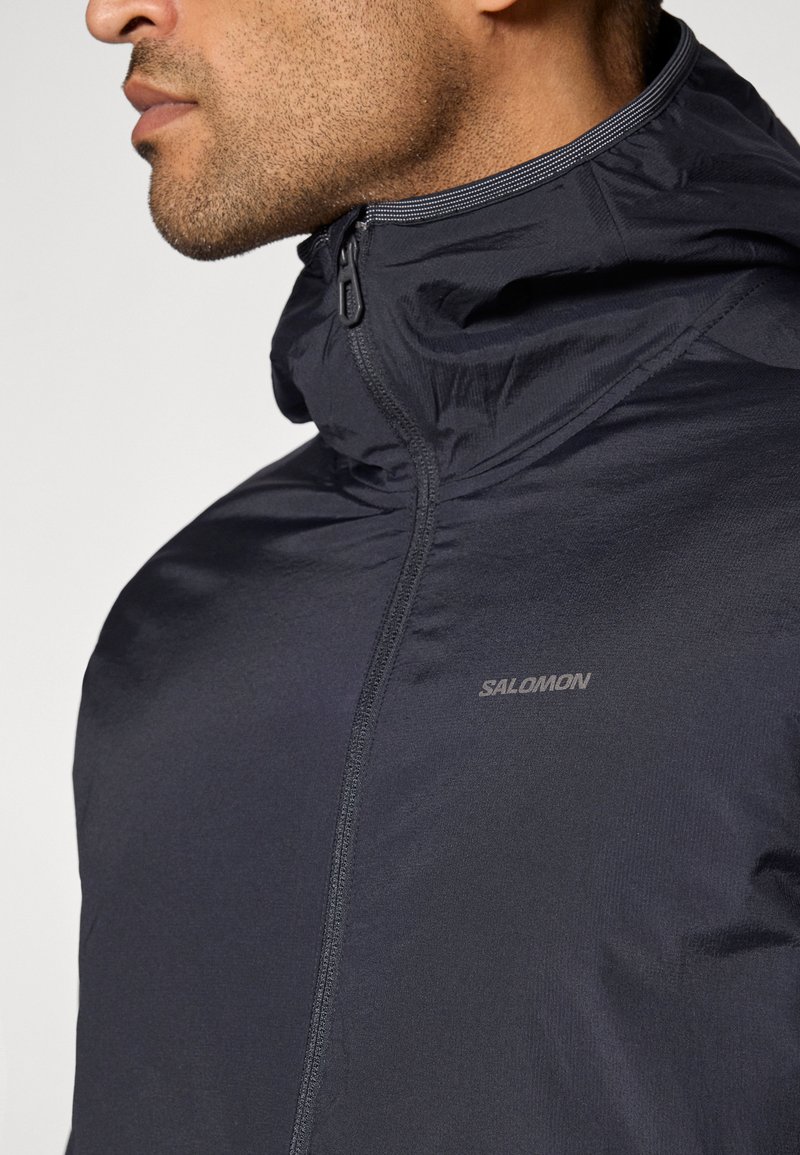 Giacca leggera nera con colletto alto e zip, con un logo discreto. Il tessuto appare liscio con una finitura opaca.